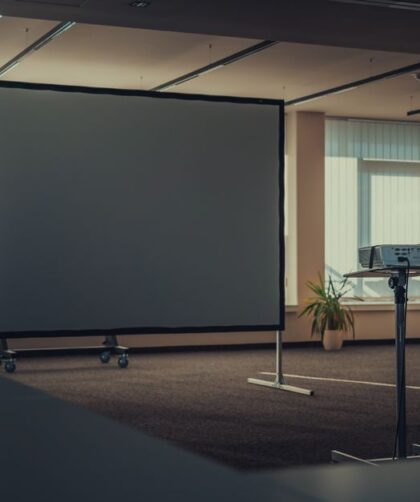 Mobilny ekran projekcyjny na kółkach oraz projektor na statywie w sali konferencyjnej